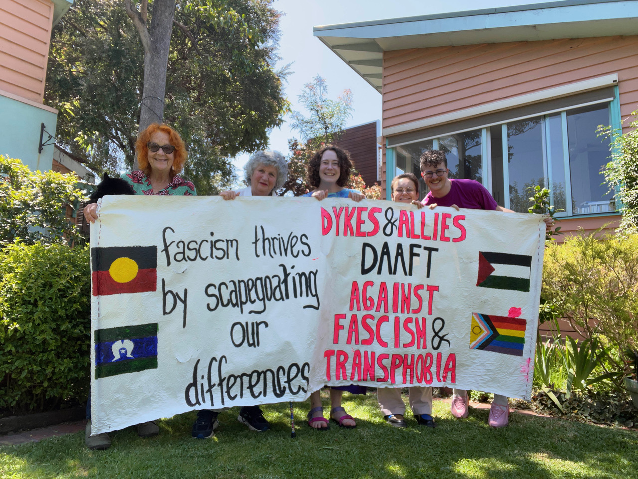 Ren Loren Britton appears on the far right of the image holding a banner with (from left to right) Di Otto, Joan Nestle, Emily Jones and Goda Klumbytė. The banner reads "fascism thrives by scapegoating our differences - DYKES & ALLIES DAAFT AGAINST FASCISM & TRANSPHOBIA". On the left there is an Australian Aboriginal Flag and an Australian Aboriginal Torres Strait Islander Flag. On the right there is a Palestinian Flag and an Intersex Inclusive Progressive Pride Flag. The group stands outside in a garden, it is green and sunny - everyone smiles and there's a cute dog, Cello in the arms of Di.